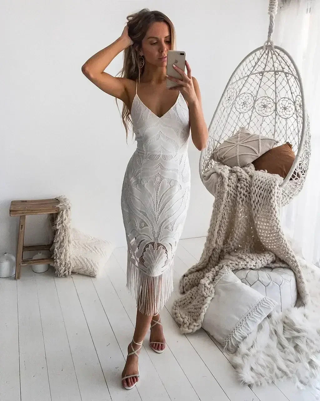 A woman in a white, intricately patterned dress takes a selfie. She stands in a softly lit room featuring a hanging chair, plush cushions, and a wooden side table.