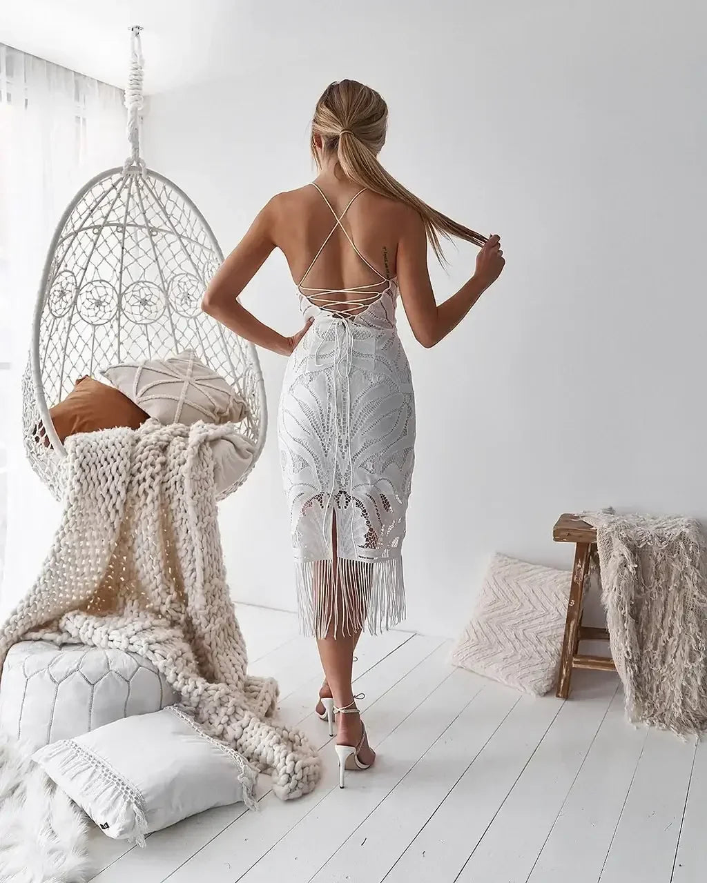 A woman is standing in a white, intricately designed dress with fringe at the hem, showcasing her back. She is in a bright room with a hanging chair and soft textiles nearby.