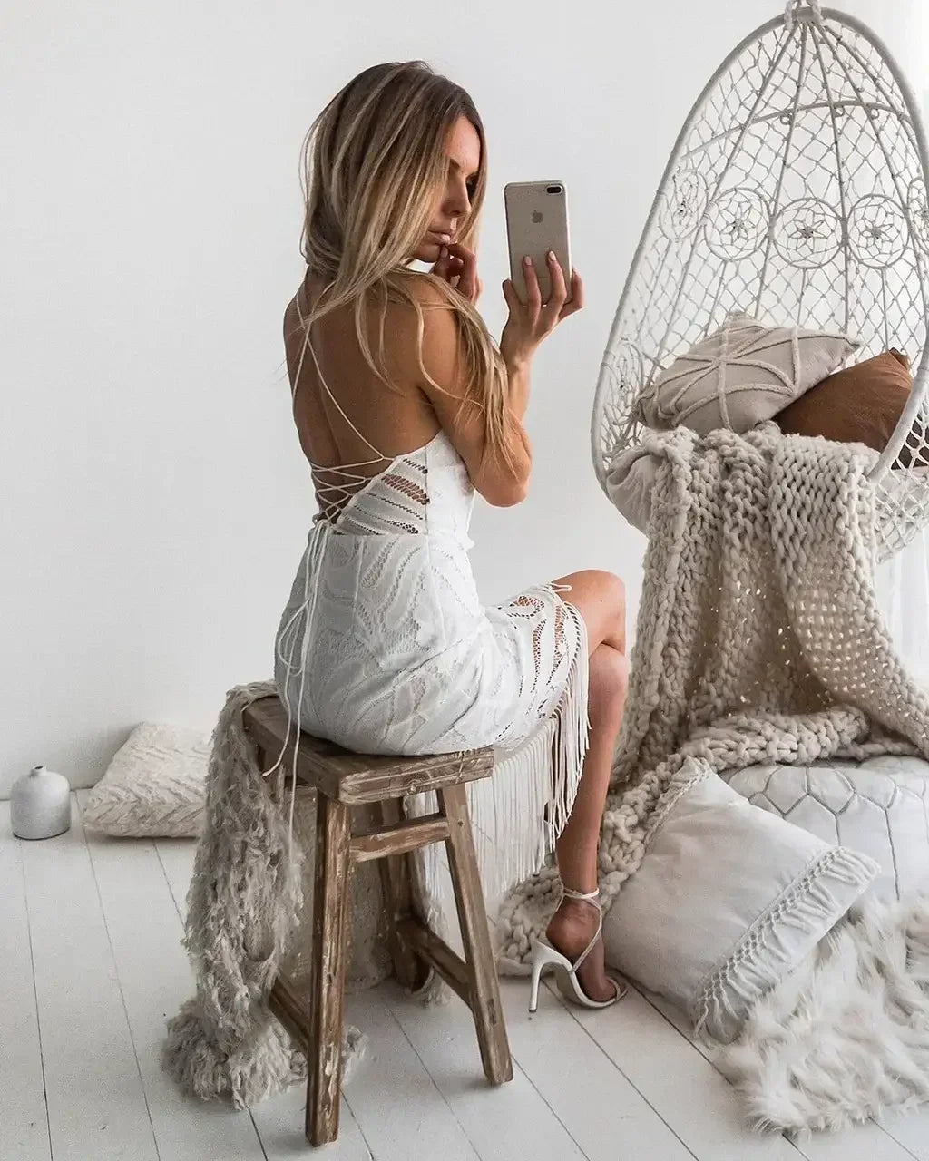 A woman in a white dress sits on a wooden stool, holding a phone and taking a selfie, surrounded by cozy, neutral-colored textiles and a woven hanging chair in a bright room.