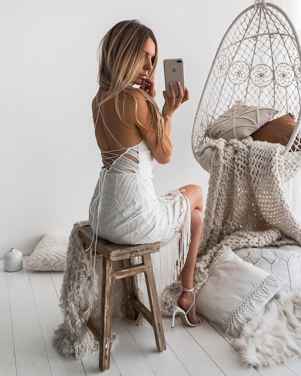 A woman in a white dress sits on a wooden stool, holding a phone and taking a selfie, surrounded by cozy, neutral-colored textiles and a woven hanging chair in a bright room.