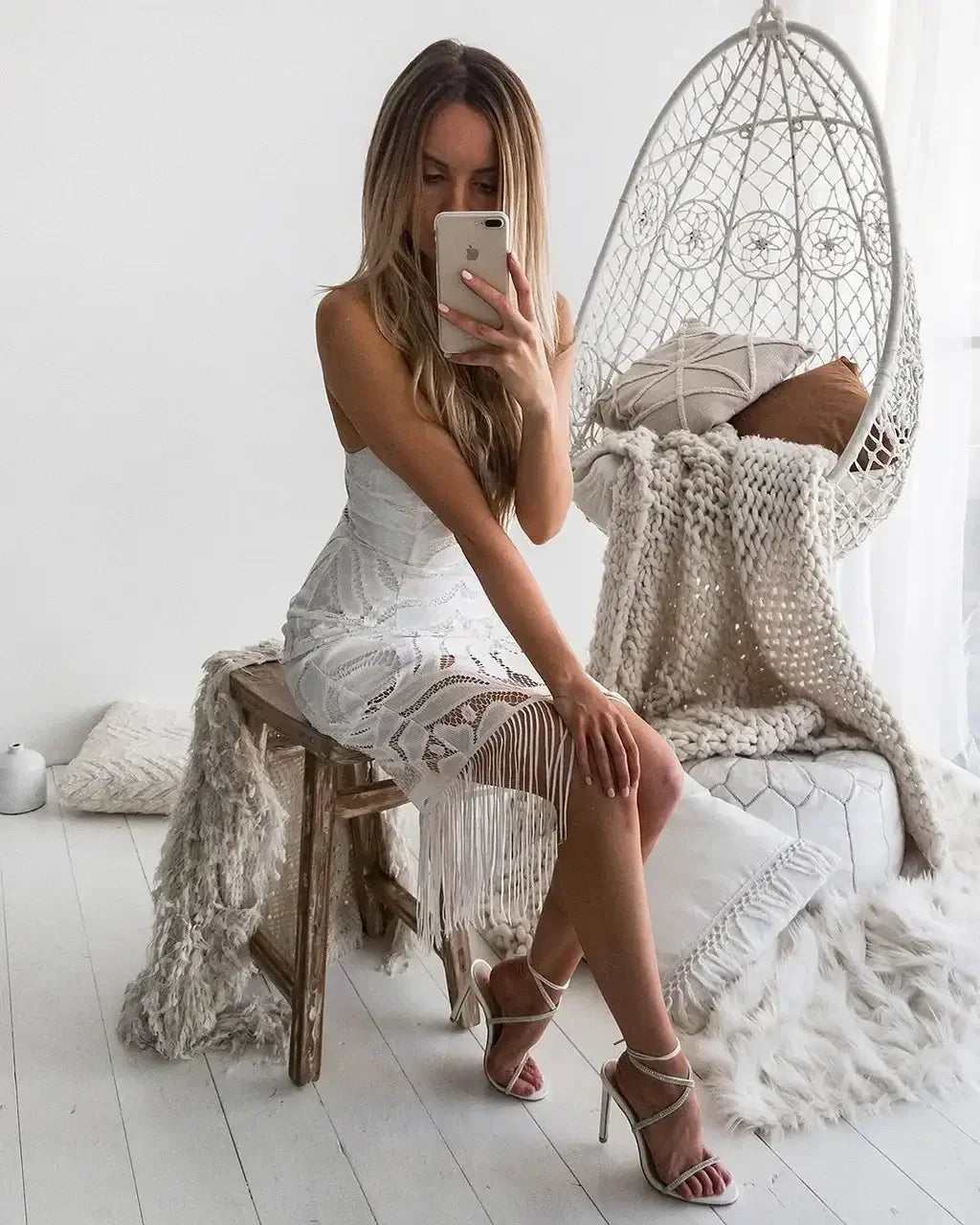A woman sits on a wooden stool, dressed in a white fringed dress, taking a selfie. She is surrounded by cozy textiles, a hanging chair, and a light, airy room.