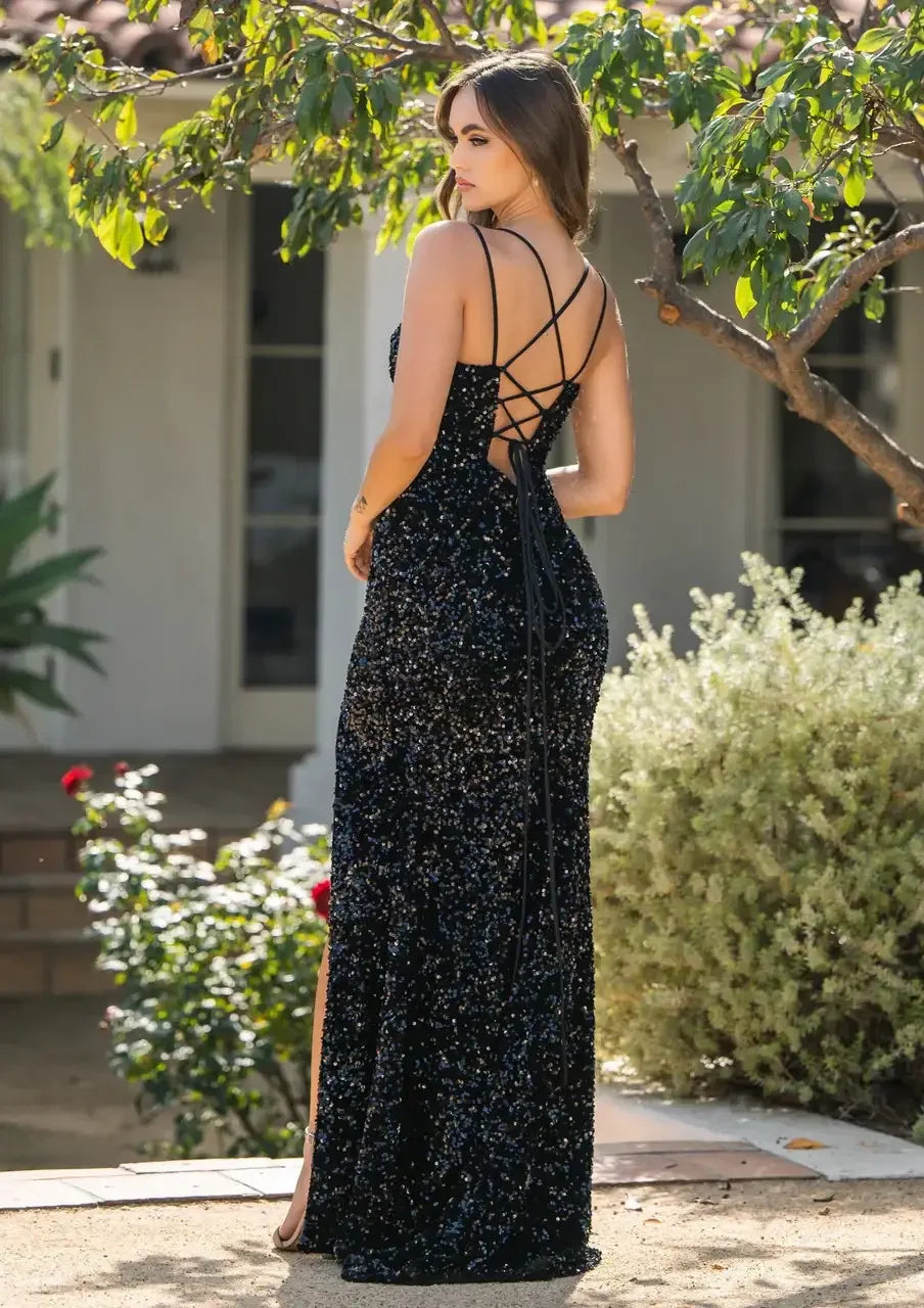 A woman wears a sequined black gown with an open back and crisscross straps, standing gracefully outdoors next to shrubs and flowers, with a house visible in the background.