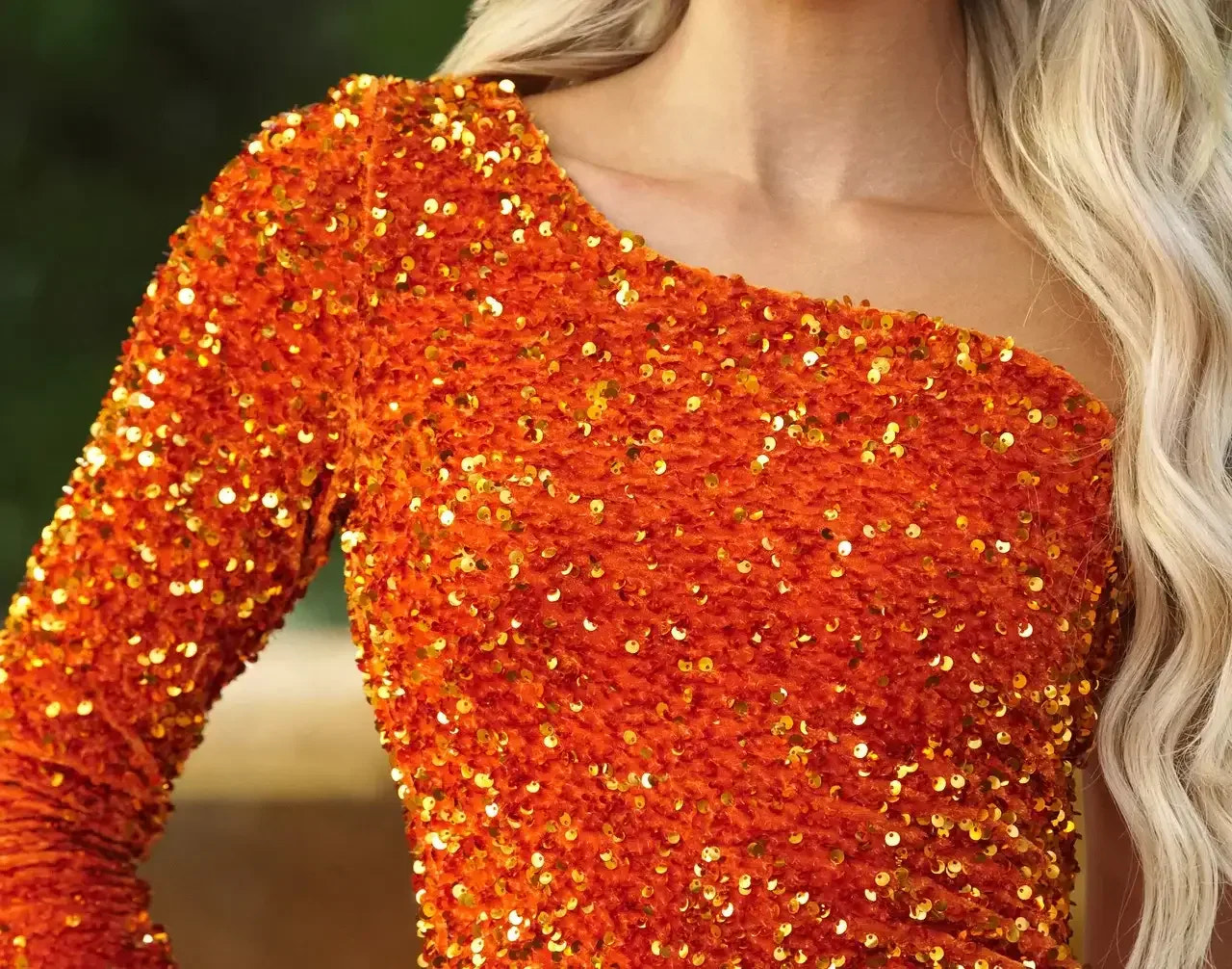 A vibrant orange dress, adorned with sparkling gold sequins, catches the light. It fits snugly on the shoulder, highlighting a textured fabric in a bright outdoor setting.