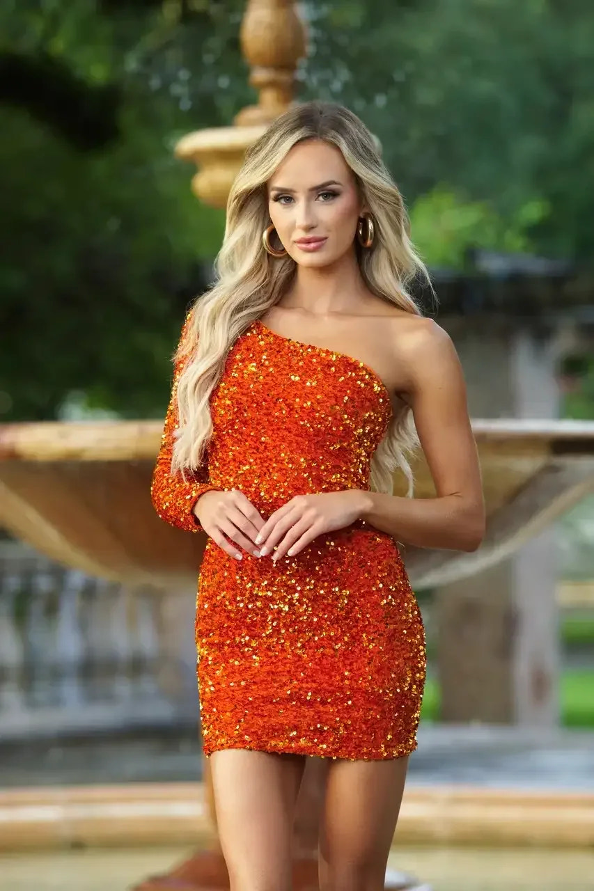 A woman stands confidently in an orange sequined dress, posing with her hands elegantly clasped. She is in a lush garden with a blurred fountain in the background.