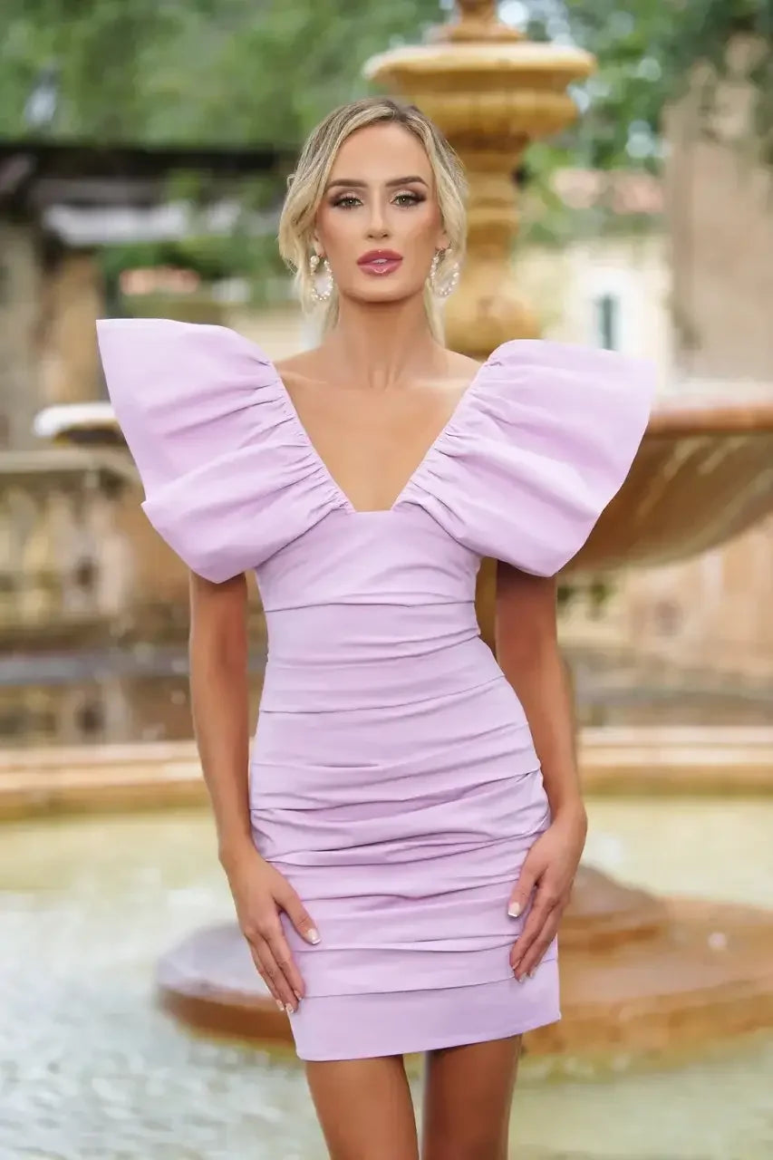 A woman stands confidently in a lilac dress with dramatic puff sleeves. She poses near a fountain surrounded by greenery, showcasing a serene outdoor environment.