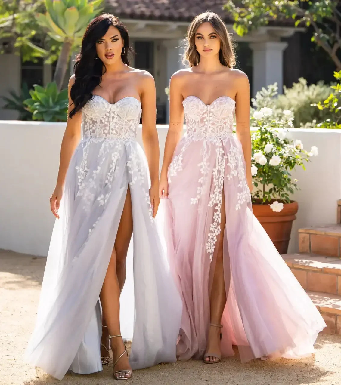 Two women stand side by side, wearing elegant, strapless dresses—one in soft gray and the other in pink. Both dresses feature floral embroidery and high slits, set against a sunlit outdoor garden.