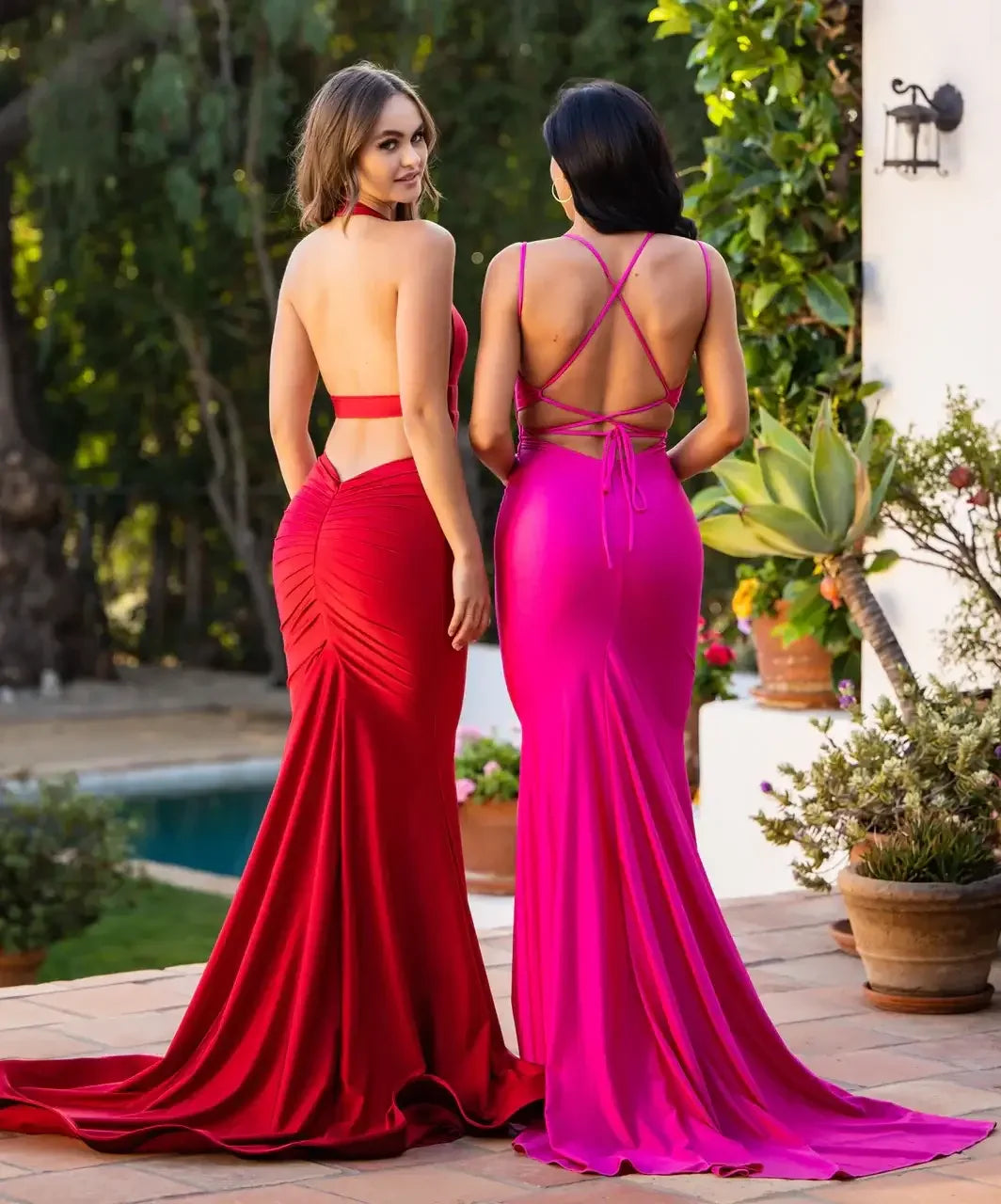 Two women stand back-to-back, wearing elegant, form-fitting gowns—one red and the other pink—with long flowing skirts. The background features greenery and a pool, enhancing the outdoor setting.