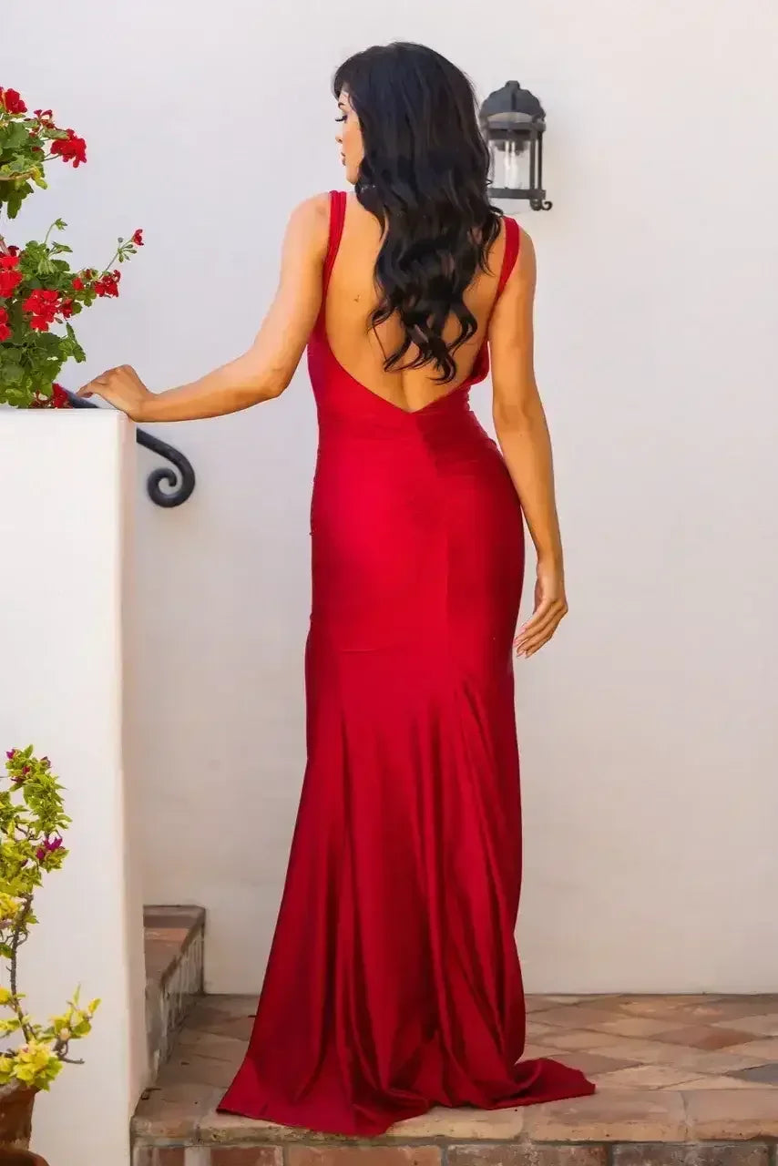 A woman in a red evening dress with a low back stands at the edge of a patio, surrounded by vibrant flowers and a stone walkway, presenting a graceful pose.