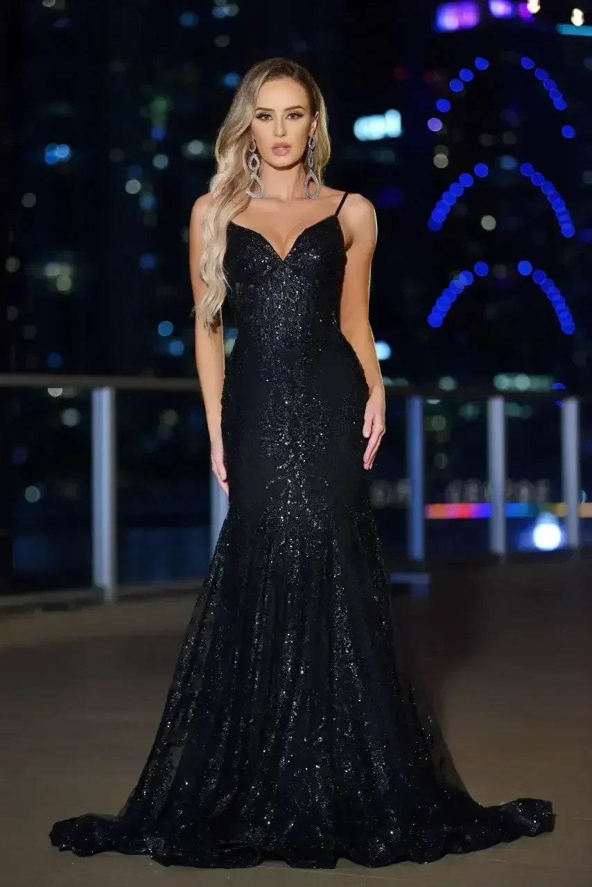 A woman stands confidently in a sparkling black gown with a fitted silhouette, posing against a blurred urban backdrop illuminated by colorful lights at night.