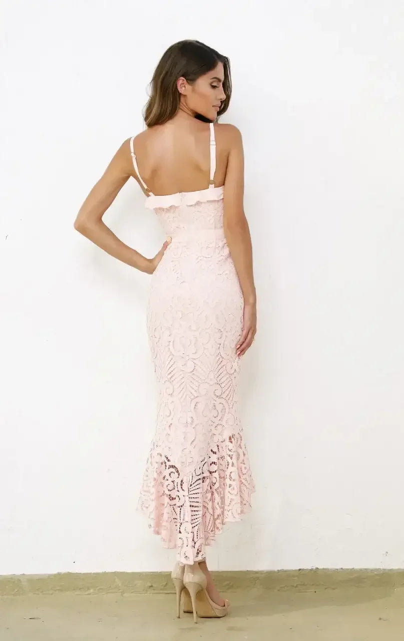 A woman is posed in a fitted pink lace dress, showcasing a flared hemline, while standing against a plain white wall with a neutral floor. She wears nude high heels.