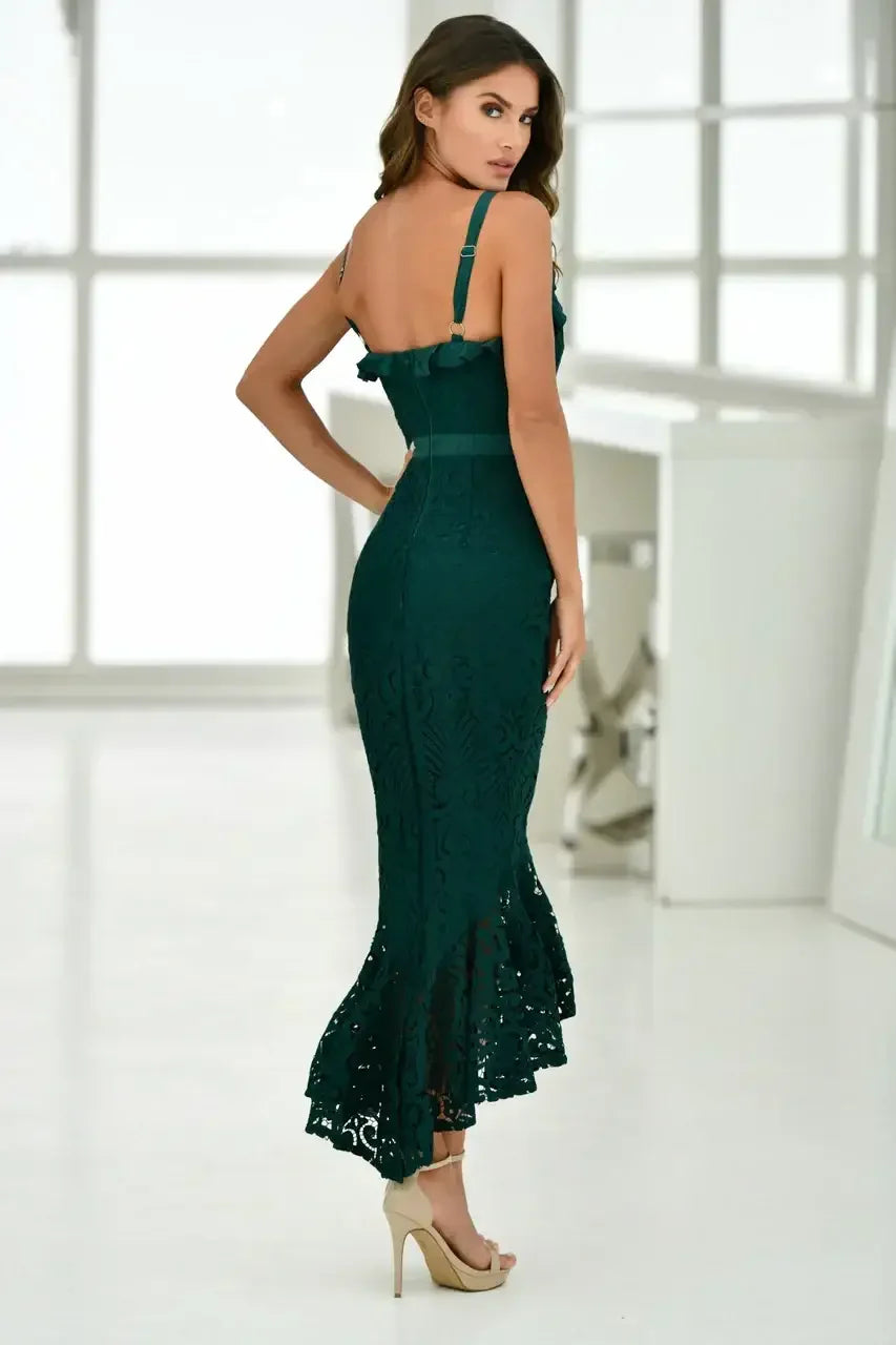 A woman stands elegantly in a form-fitting, dark green dress with intricate lace patterns and a flared hem. She is in a bright, modern room with large windows.