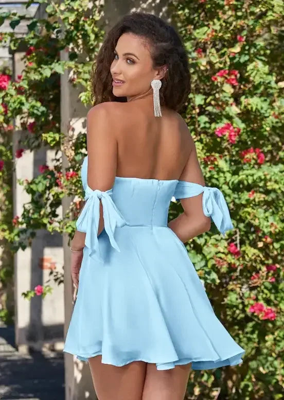 A woman in a light blue, off-the-shoulder dress stands with her back to the viewer, smiling over her shoulder. She is surrounded by lush greenery and blooming flowers.