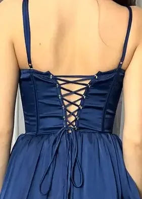 A navy blue dress features a lace-up back with crisscrossed straps and small decorative beads, positioned on a person viewed from behind in a softly lit indoor setting.