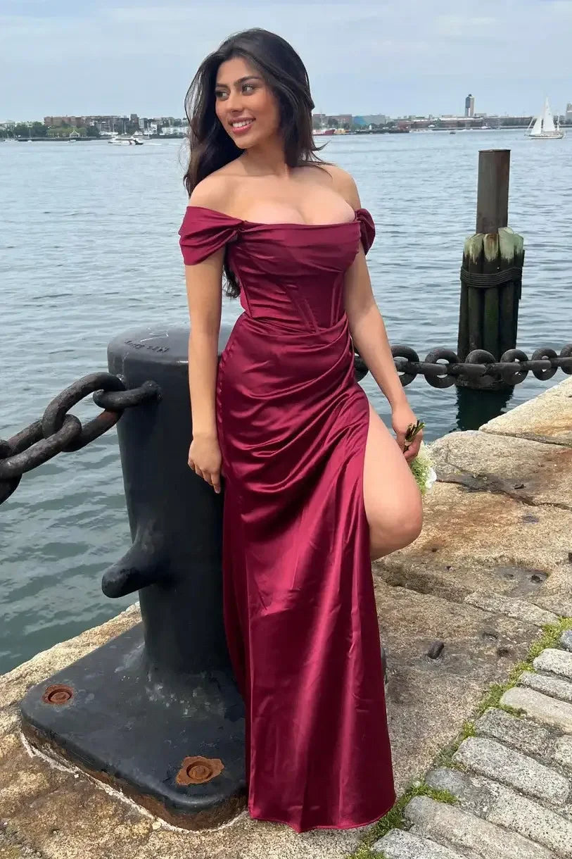 A woman in a burgundy off-shoulder dress stands beside a large mooring post along a waterfront, smiling, with a chain and rocky pavement visible, and boats in the background.