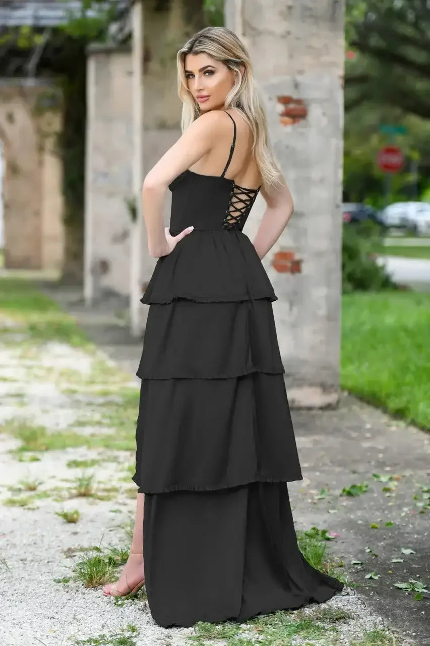 A woman stands in a flowing black dress with layered ruffles, adjusting her hair. The setting features weathered stone walls and greenery, conveying a serene outdoor atmosphere.