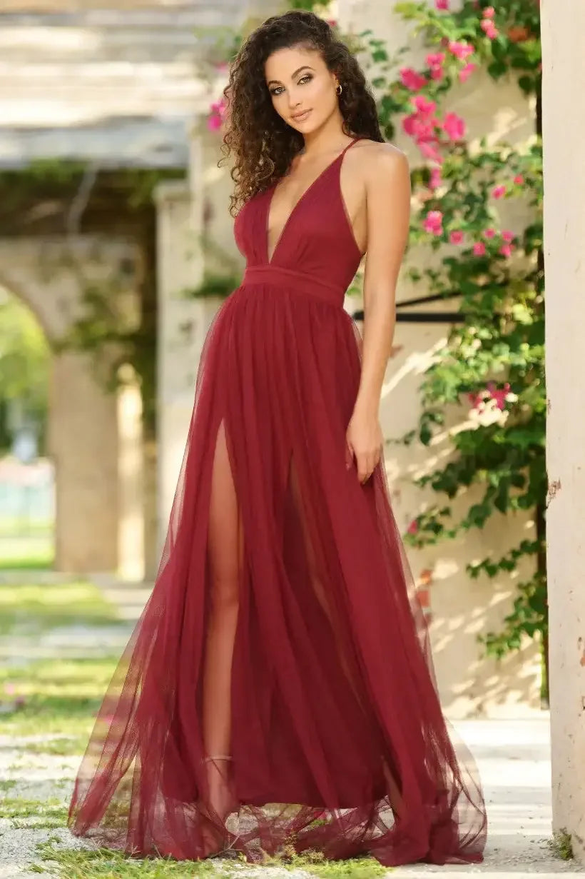 A woman stands gracefully in a flowing burgundy dress with high slits, smiling radiantly. She is surrounded by lush greenery and pink flowers against a rustic backdrop.