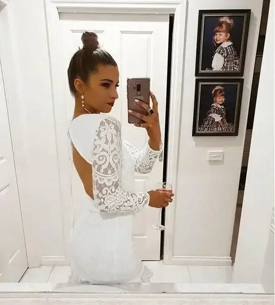 A woman in a white lace dress poses for a selfie in front of a mirror, holding a glass. Behind her, framed photos of children adorn a white wall.