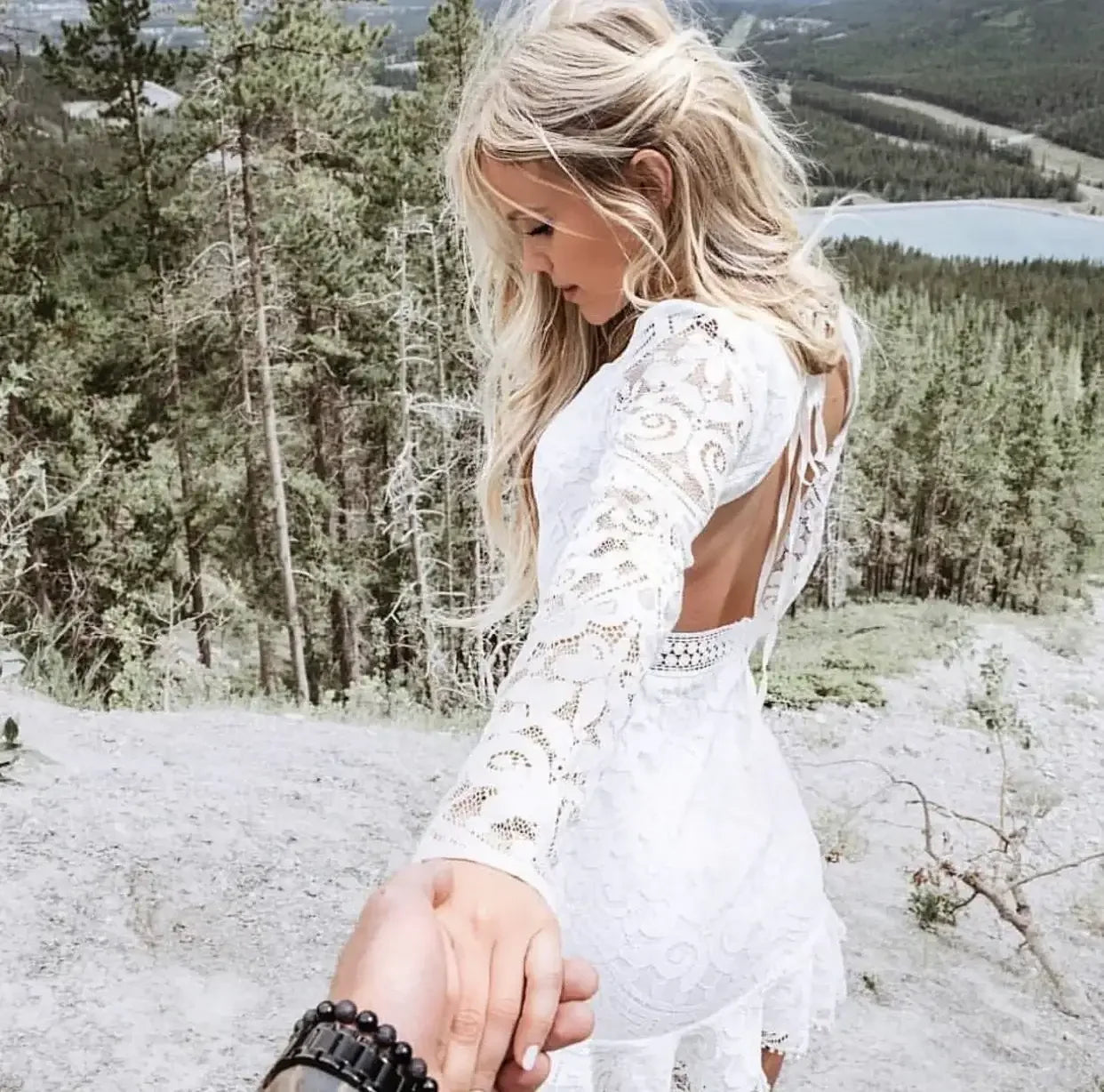 A woman in a white lace dress holds hands with a hidden person while looking back, standing on a rocky hillside surrounded by trees and a view of a distant lake.