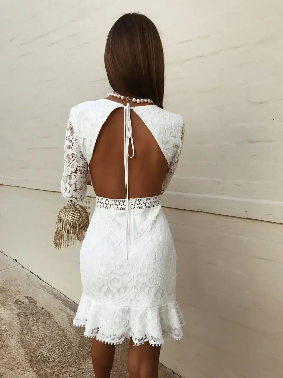 A woman stands with her back to the viewer, wearing a white lace dress with an open back, fringed handbag in hand, against a smooth light-colored wall.