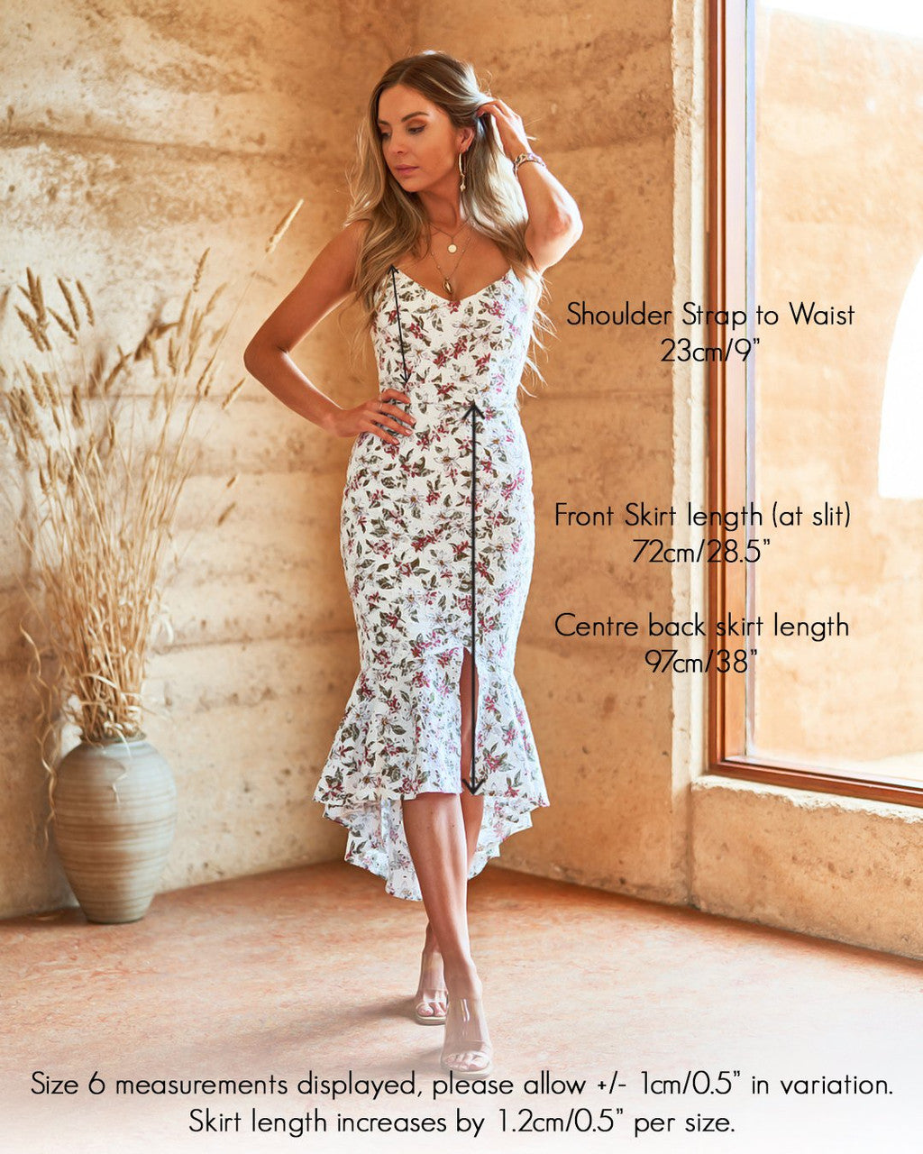 A woman in a floral dress stands gracefully, adjusting her hair. She is inside a textured, earthy-toned room with a large window and a plant nearby. Text details skirt measurements and variations.