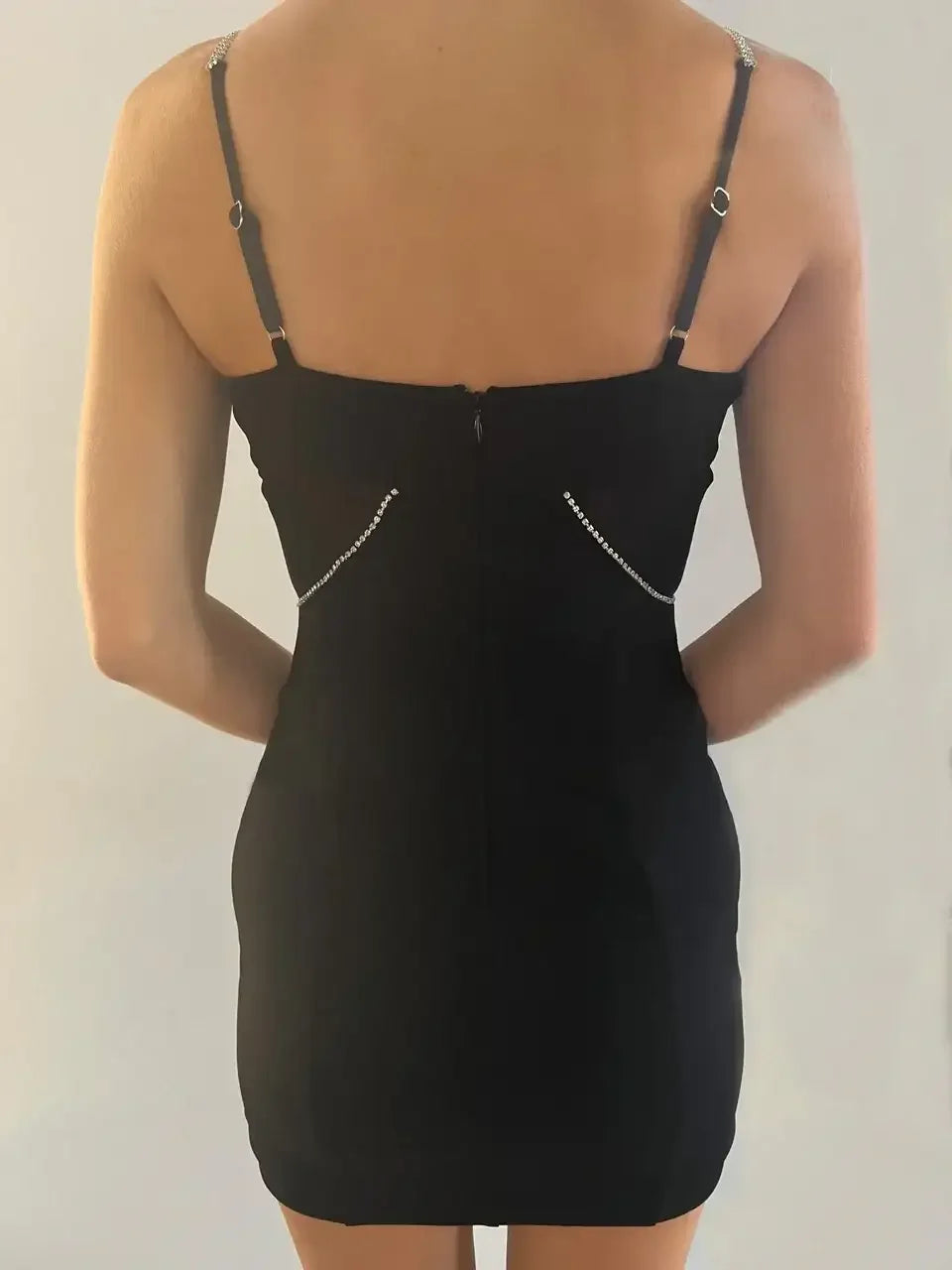 A person is standing with their back facing the viewer, wearing a fitted black dress with thin straps. The dress features decorative rhinestones on the sides, against a light background.