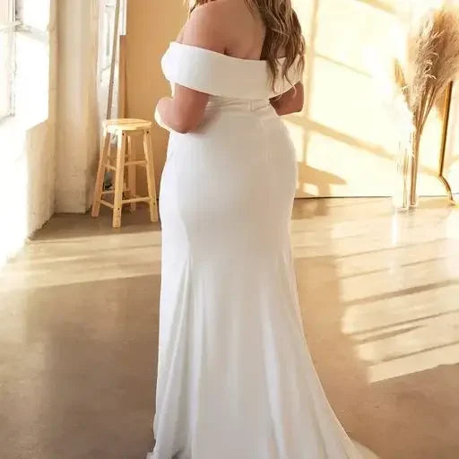 A person in a flowing white dress stands with their back to the viewer, gently resting their hands on their shoulders, illuminated by soft natural light in a bright, airy room with a wooden stool nearby.