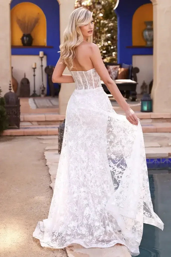 A woman in a flowing white gown stands gracefully with her back to the viewer, showcasing intricate lace details. She is set in a picturesque outdoor environment with decorative elements.