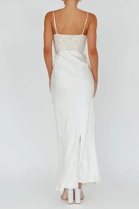 A woman stands in a white, elegant, floor-length dress with lace detailing on the bodice. The backdrop is plain, emphasizing her outfit and posture. She wears silver heels.