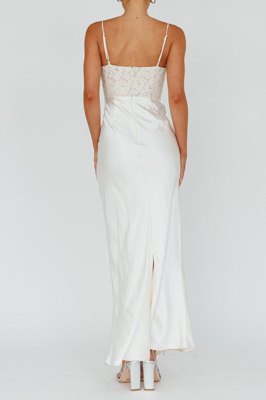 A woman stands in a white, elegant, floor-length dress with lace detailing on the bodice. The backdrop is plain, emphasizing her outfit and posture. She wears silver heels.