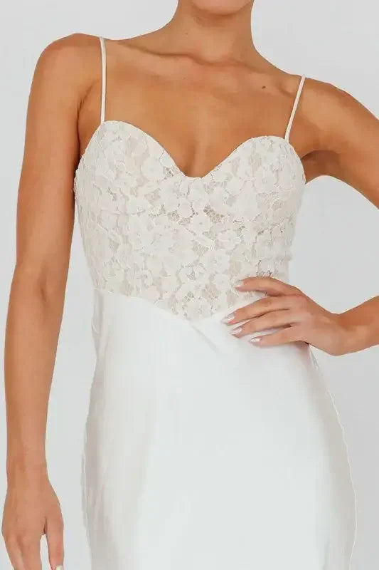 A woman wears a white dress featuring a lace bodice and thin straps, showcasing elegance while her hand rests gently on her hip, set against a plain background.