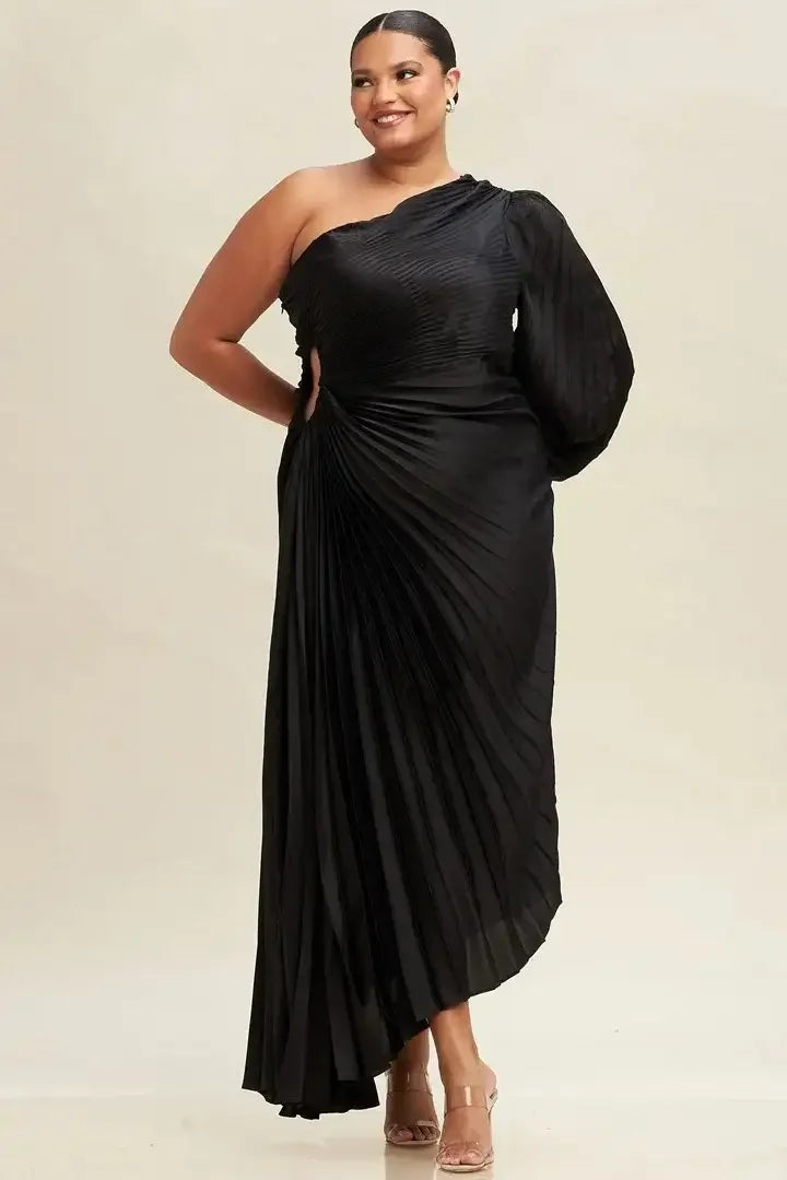 A woman stands confidently in a pleated, asymmetrical black dress, smiling. The dress has one long sleeve and a stylish cutout. The background is a soft, light color.