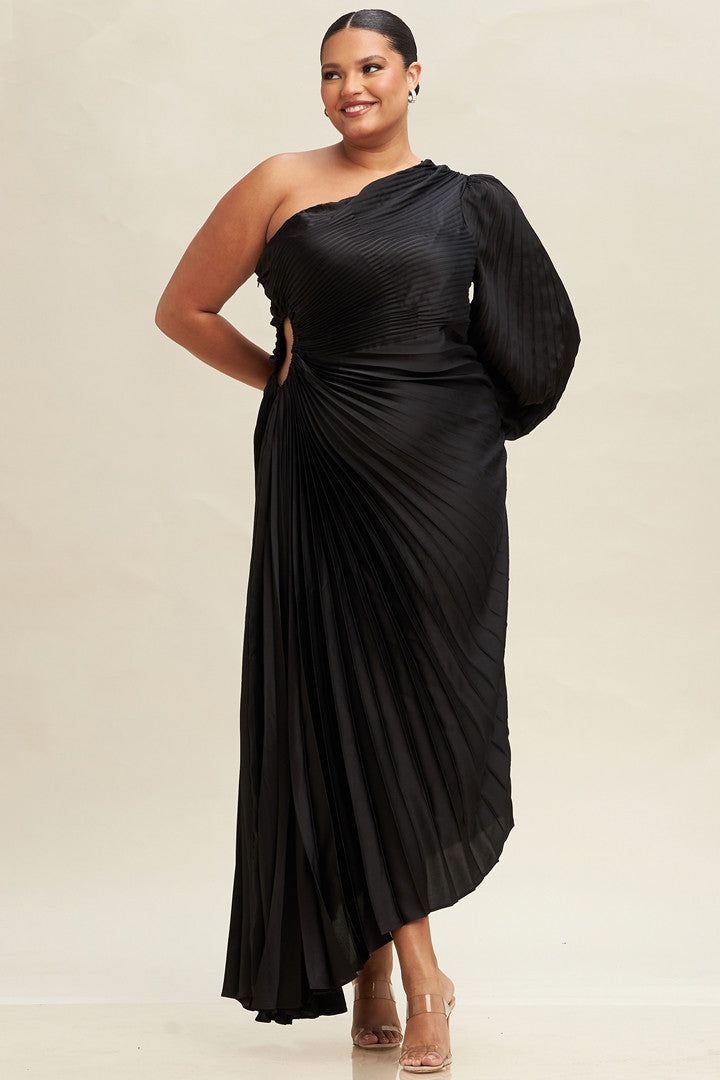 A woman stands confidently in a pleated, asymmetrical black dress, smiling. The dress has one long sleeve and a stylish cutout. The background is a soft, light color.