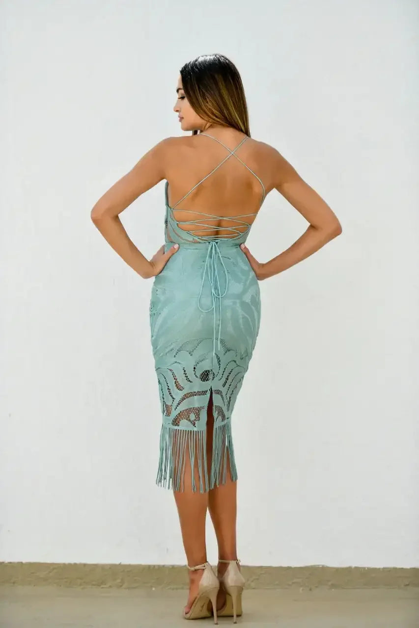 A woman models a fitted mint green dress with intricate patterns and fringe detailing. She stands against a plain white wall, showcasing the dress's back and spaghetti strap design.