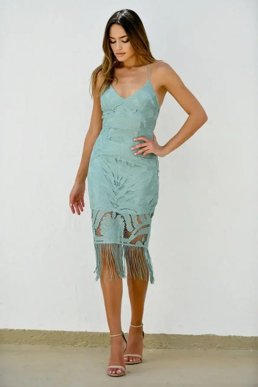 A woman is standing gracefully in a light teal, fringed dress, looking down thoughtfully. She has long hair and strappy heels, with a textured white wall behind her.