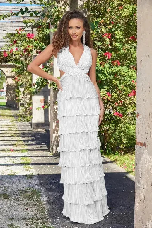 A woman stands elegantly in a tiered white dress, surrounded by blooming flowers and greenery in a picturesque outdoor setting, conveying a sense of grace and tranquility.