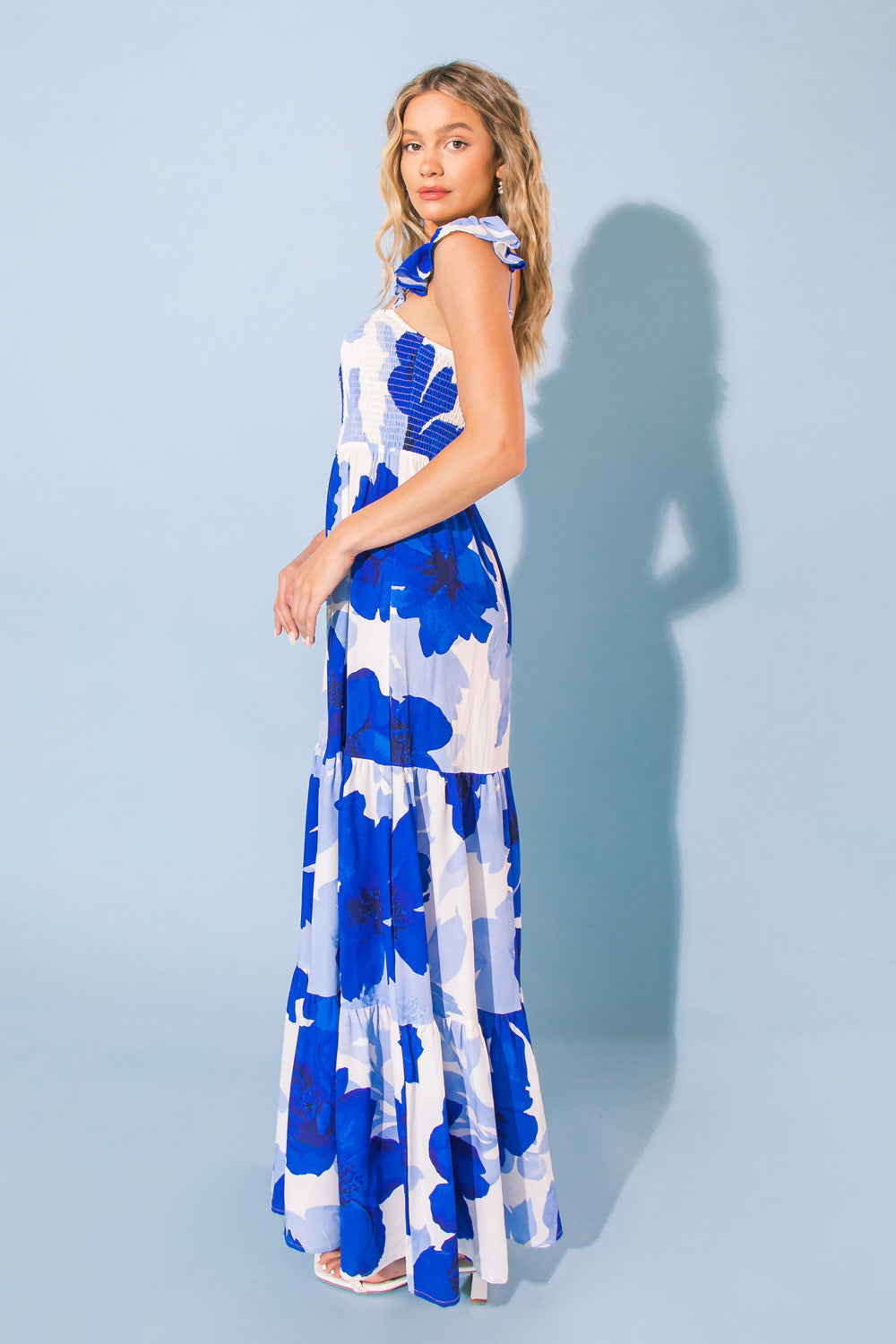 A woman stands sideways, showcasing a long, floral dress with blue and white patterns. She poses against a light blue background, her hair cascading in loose waves.