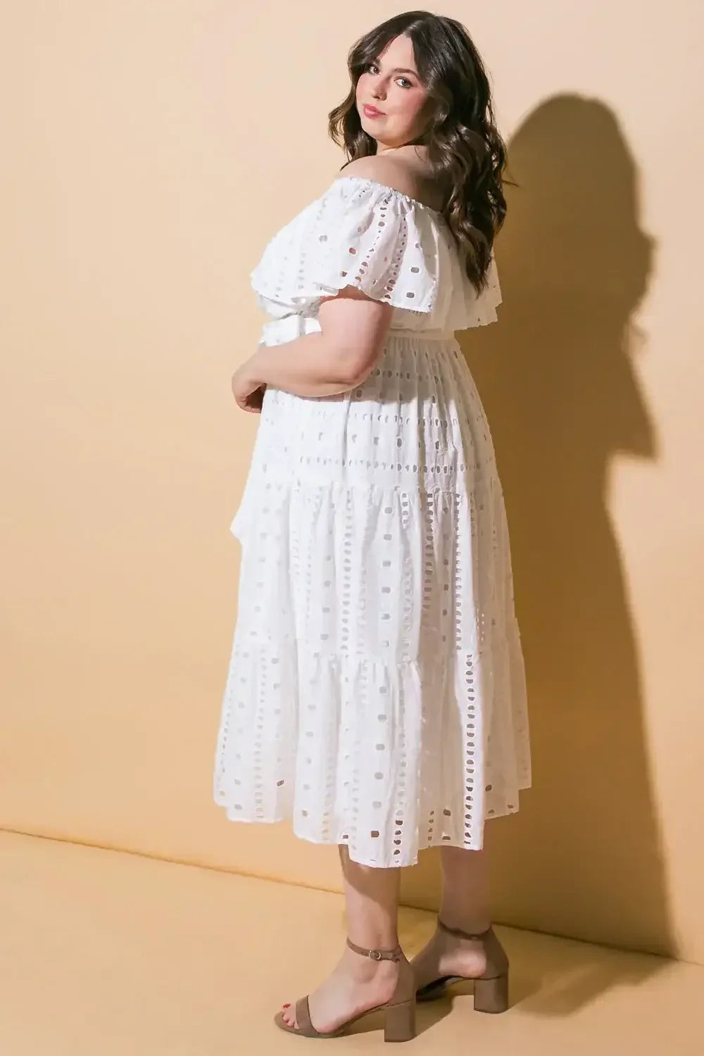 A woman stands sideways, wearing a flowing white off-the-shoulder dress with eyelet patterns. She has long, wavy hair and is positioned against a soft beige background.