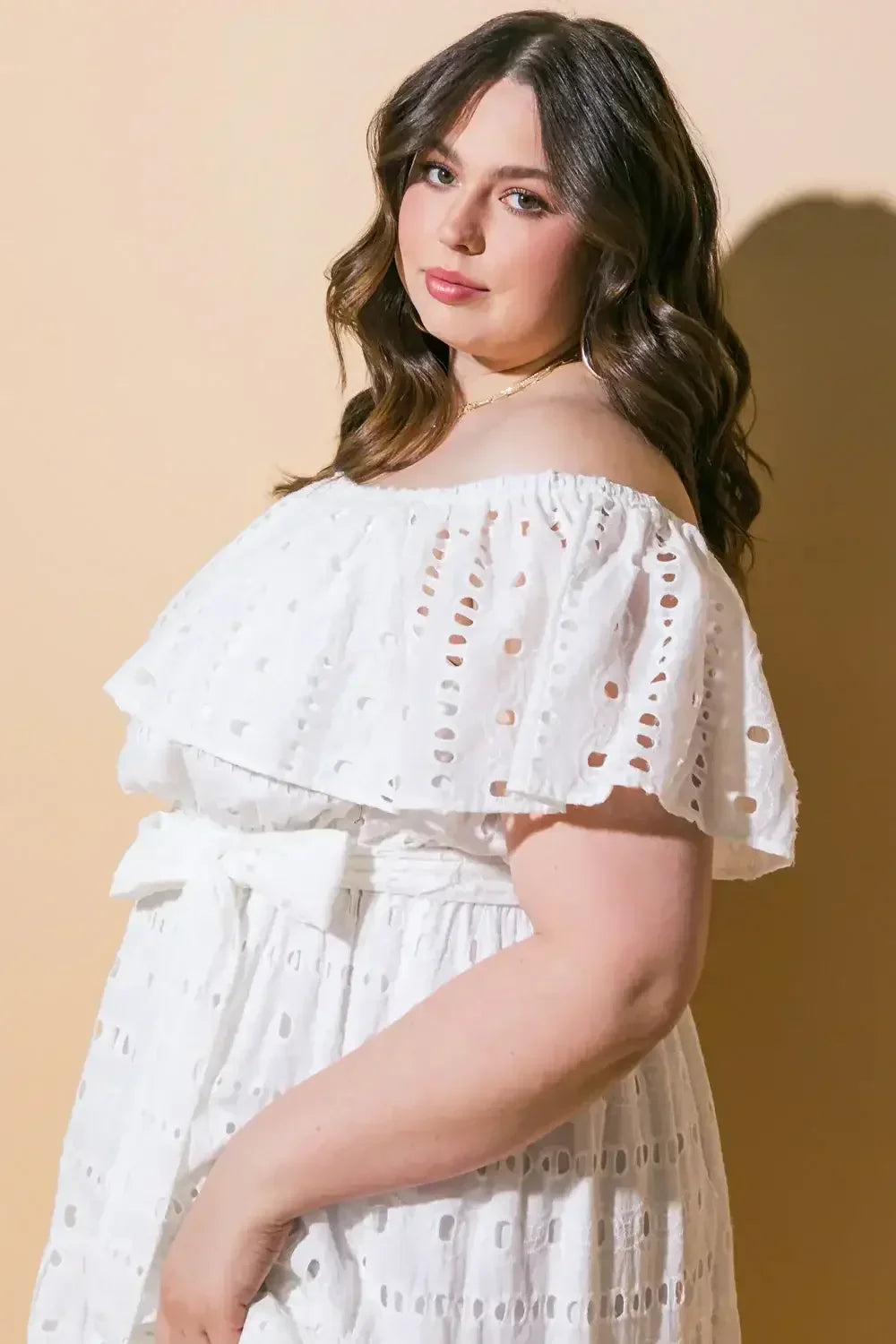 A woman in an off-the-shoulder white dress with eyelet details stands confidently, facing slightly sideways. She has long, wavy hair and a soft expression, set against a light brown background.