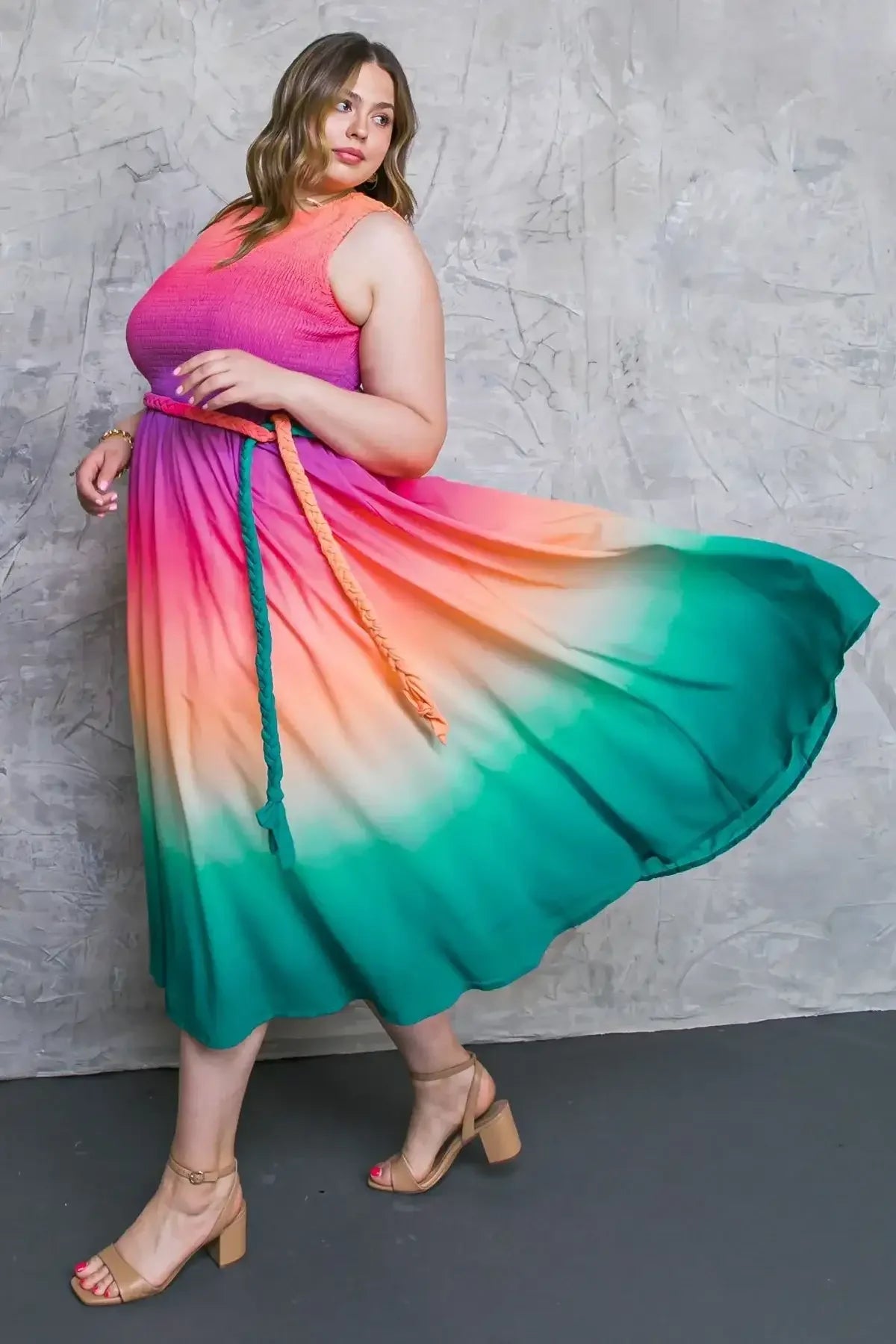A woman in a vibrant, multi-colored dress spins gracefully, the fabric flowing outward. She wears a braided belt and strappy heeled sandals, posing against a textured gray background.