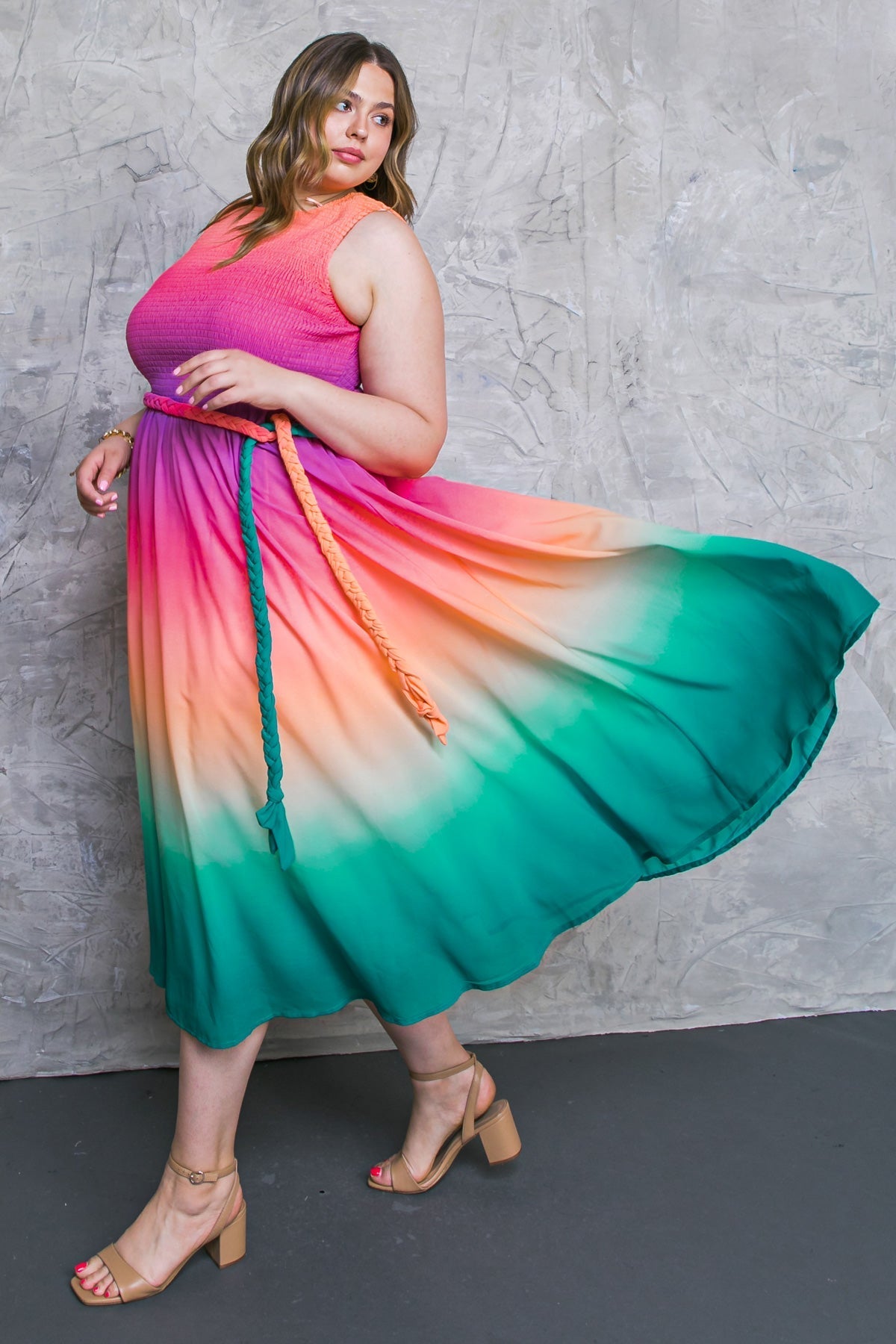 A woman in a vibrant, multi-colored dress spins gracefully, the fabric flowing outward. She wears a braided belt and strappy heeled sandals, posing against a textured gray background.
