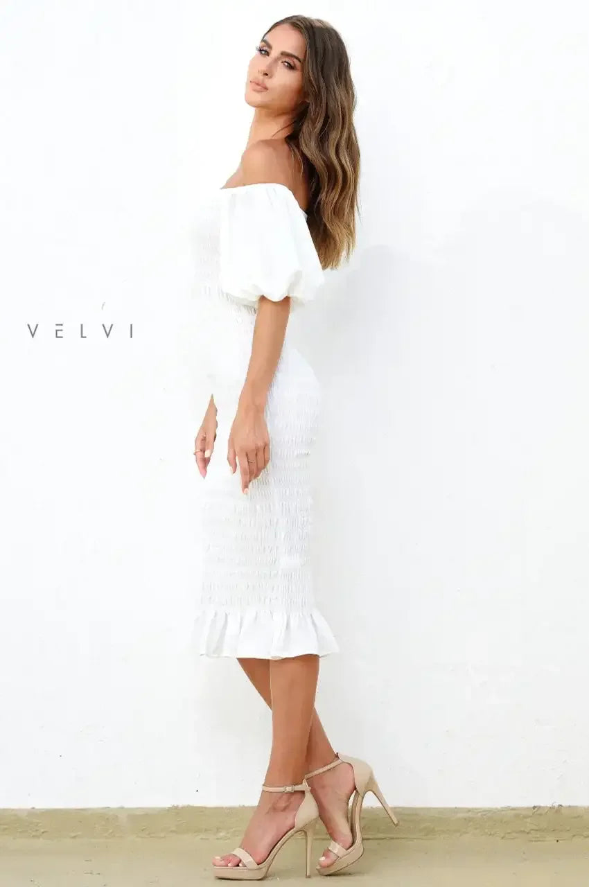 A woman wearing a fitted white dress with puffed sleeves poses sideways. She stands on a light-colored floor against a textured white wall. The setting appears bright and clean.