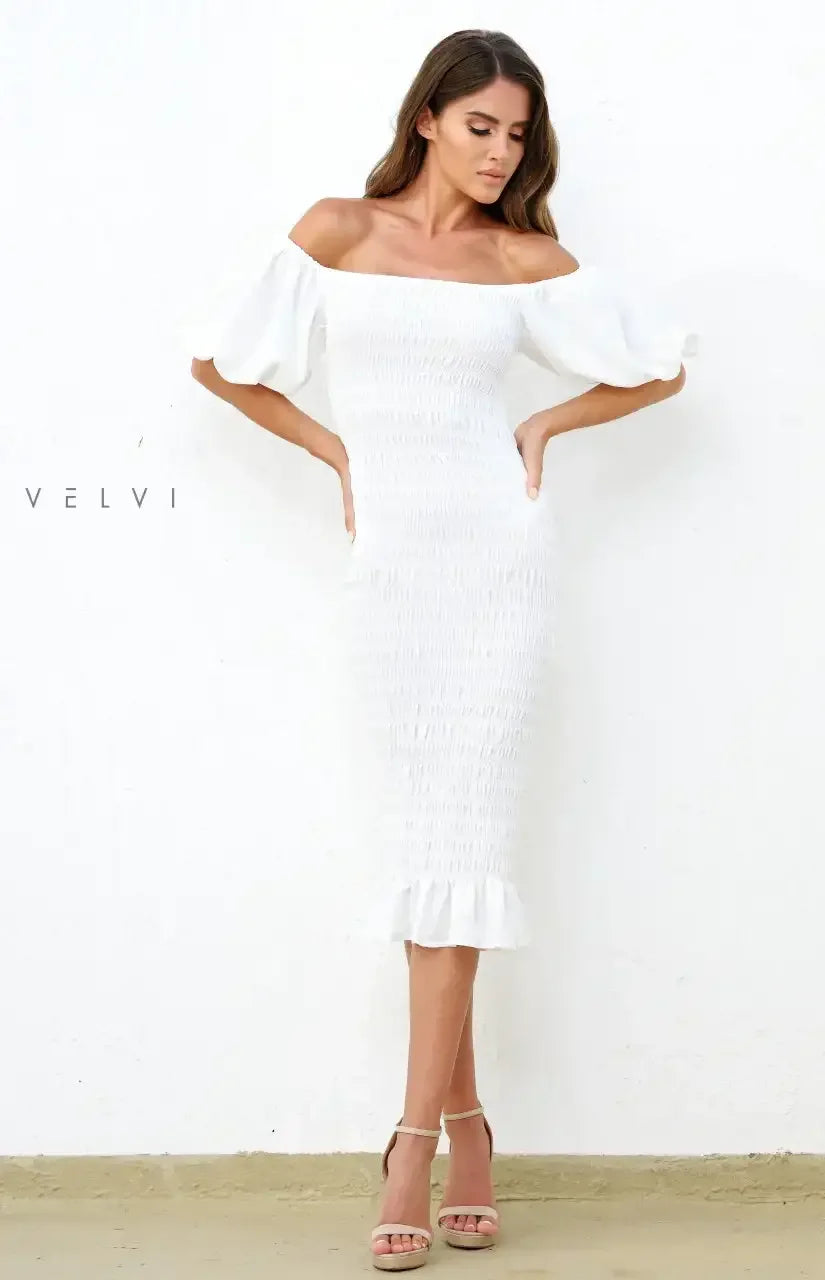 A woman poses in a fitted white off-the-shoulder dress with puffed sleeves, standing against a textured white wall, highlighting her elegant style. She wears nude high-heeled sandals.