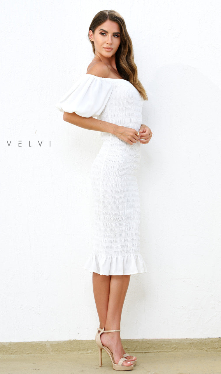 A woman stands sideways, wearing a fitted white off-shoulder dress with a ruffled hem. The background is plain white, enhancing her elegant look, complemented by nude high heels.