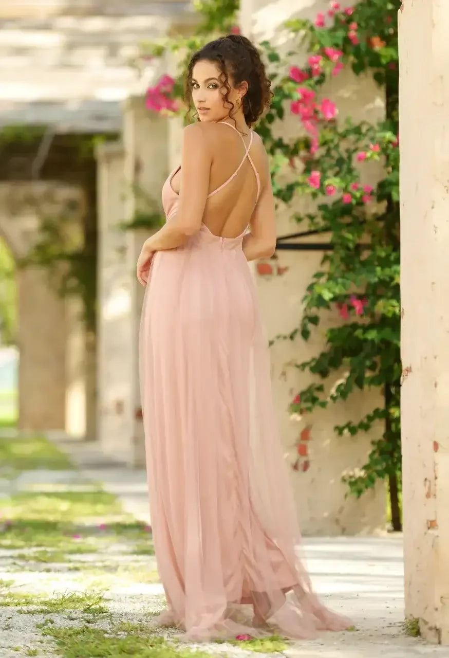 A woman in a sleeveless, light pink gown stands with her back to the viewer, surrounded by greenery and brick walls, with pink flowers blooming nearby.