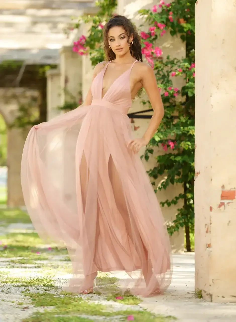 A woman in a flowing, light pink dress stands gracefully, with one arm extending the fabric. She is surrounded by lush greenery and blooming flowers in an outdoor setting, enhancing a serene atmosphere.