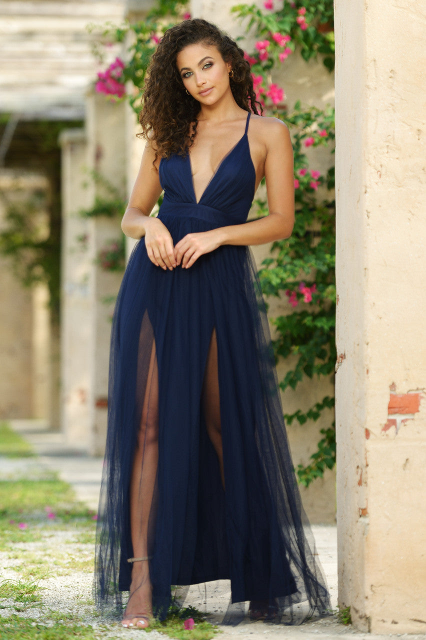 A woman stands elegantly in a flowing navy gown with high slits, framed by blossoming flowers and rustic walls in a serene outdoor setting. Sunlight highlights her curly hair and features.