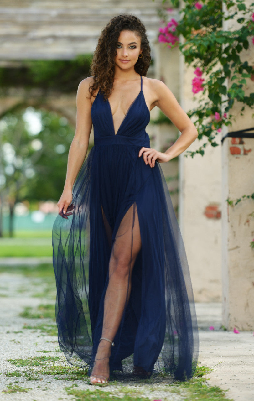 A woman wears a flowing, navy blue gown with a deep V-neck and thigh-high splits, confidently standing in a lush, outdoor setting decorated with pink flowers and greenery.