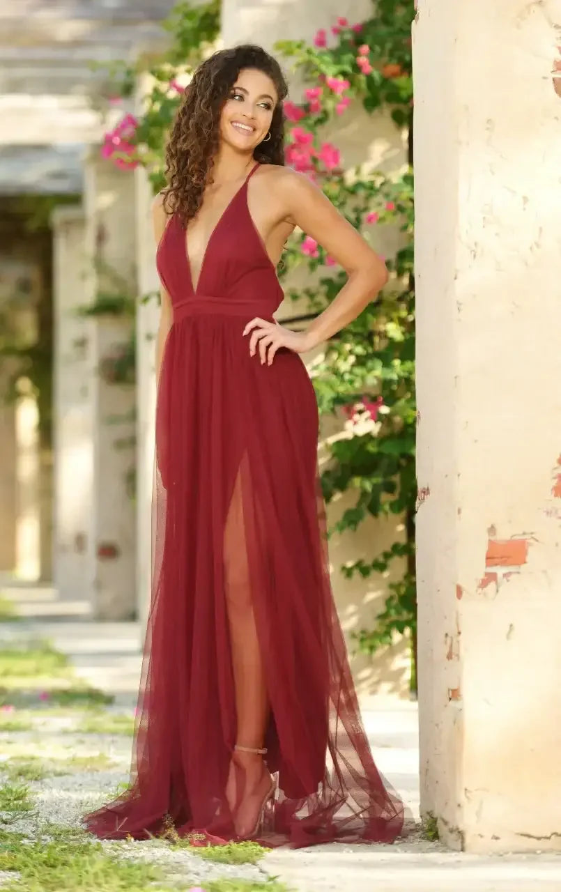 A woman in a long burgundy dress, with a plunging neckline, poses confidently outdoors. She stands near weathered stone columns with vibrant flowers in the background, showcasing a sunny, lush environment.