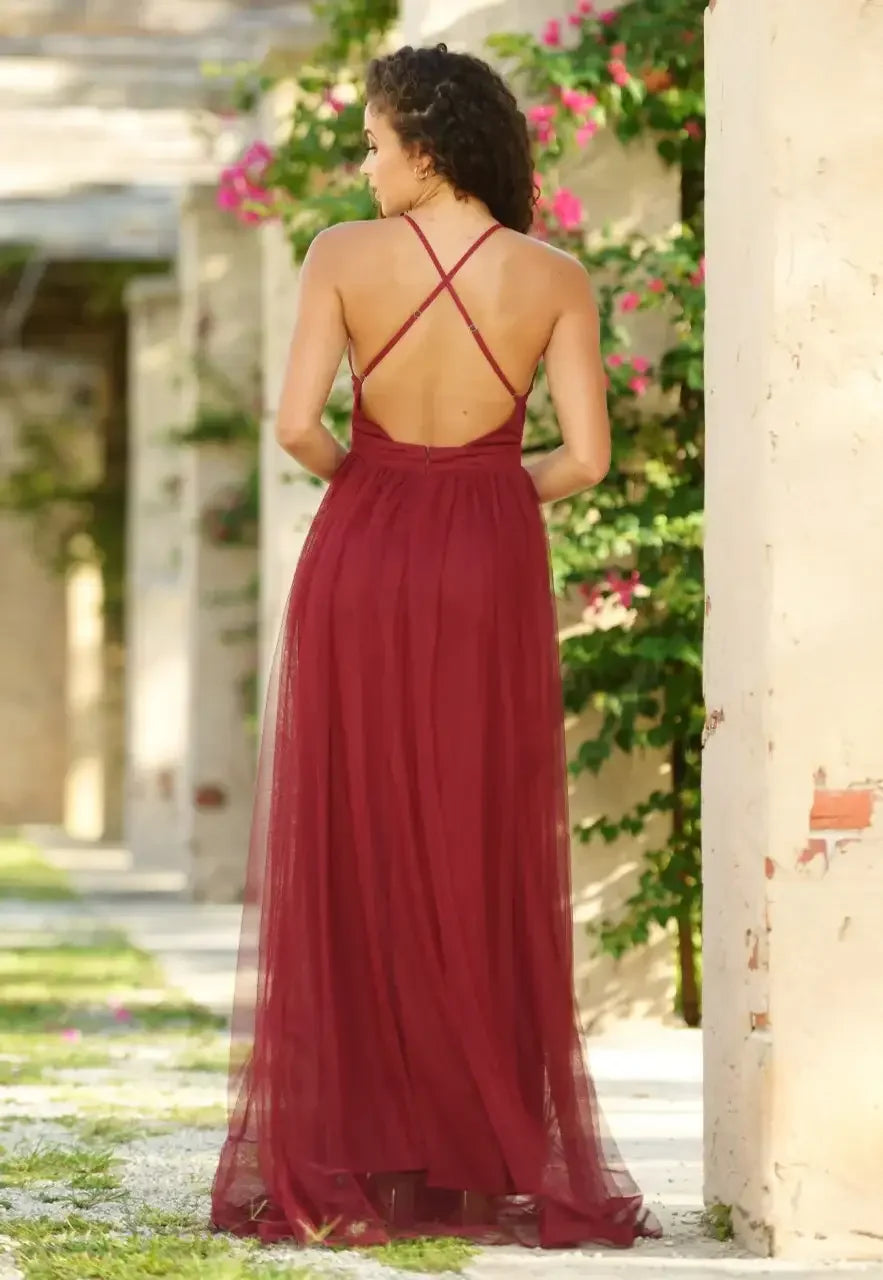 A woman in a backless, burgundy dress stands with her back turned, surrounded by a lush outdoor setting featuring greenery and architectural elements in soft focus.