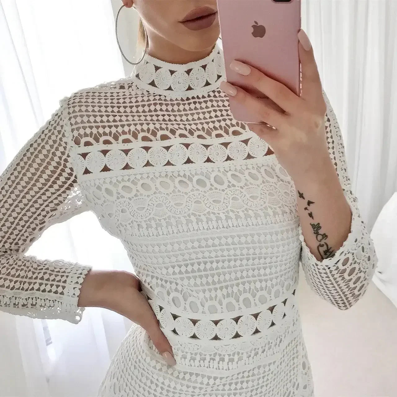 A person wears an intricate white lace dress and poses while taking a selfie with a pink phone. The background features soft white curtains, enhancing a bright, stylish atmosphere.